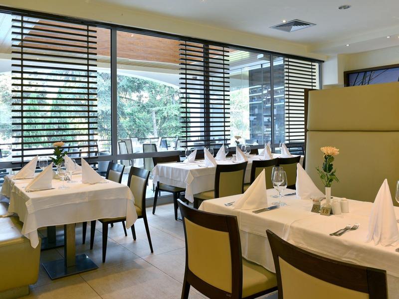 Světle zařízená jídelna s prostřenými stoly a velkými okny s dřevěnými žaluziemi.