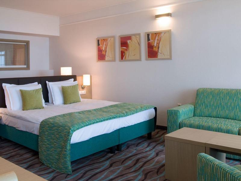 Moderní hotelový pokoj s manželskou postelí, zelenou pohovkou a abstraktní nástěnnou dekorací.