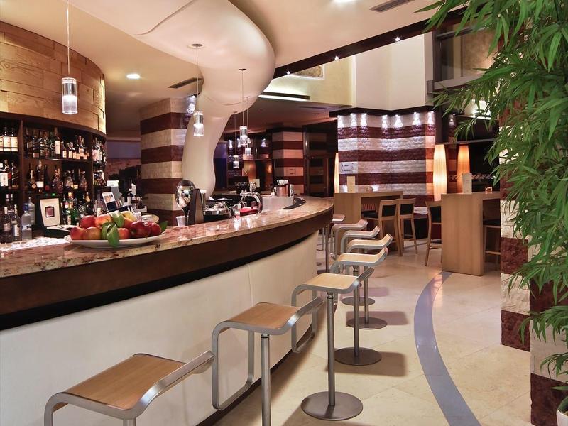 Moderní hotelový bar s barovými židlemi, elegantním osvětlením a dekorativní bambusovou rostlinou.