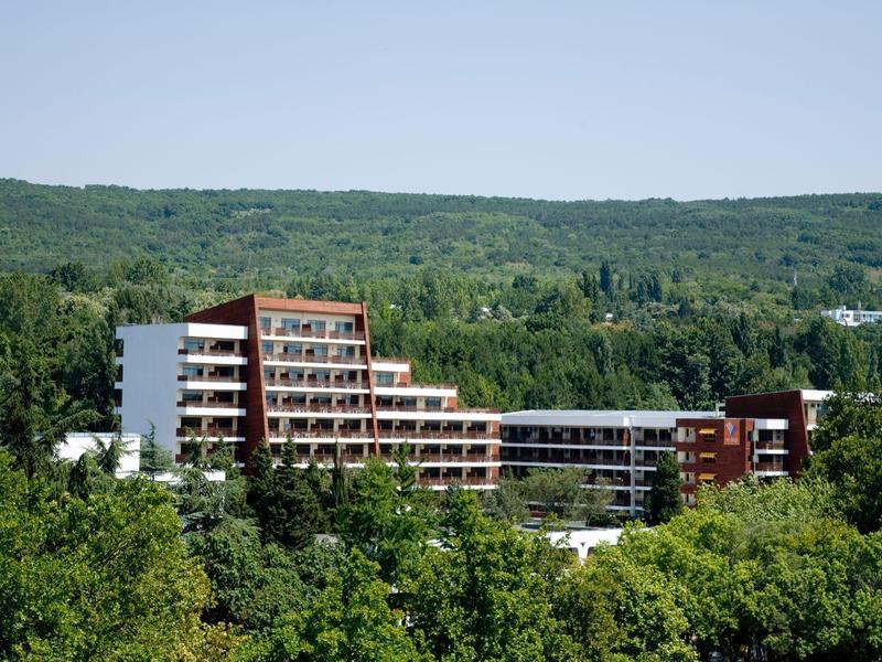 Moderní hotelová budova s několika patry obklopená zelenou krajinou a zalesněnými kopci.