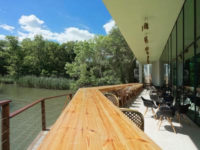 Taras z drewnianą balustradą i stolikami nad brzegiem rzeki obok nowoczesnego budynku.