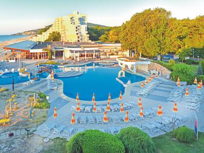 Espace piscine d'hôtel avec parasols et palmiers, entouré de bâtiments et vue sur la mer.