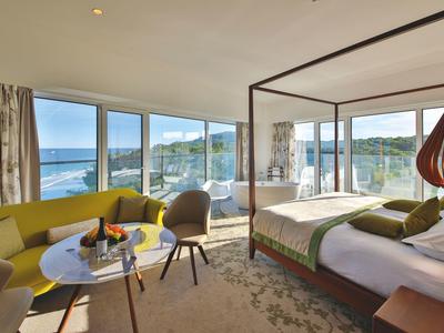 Nowoczesny pokój hotelowy z łóżkiem podwójnym, żółtą sofą i dużymi oknami z widokiem na morze.