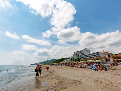 Piaszczysta plaża z ludźmi w wodzie i na brzegu, hotele i parasole w tle.