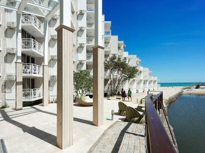 Bâtiment hôtelier blanc moderne au bord de l'eau avec balcons et coin salon sous un ciel clair.