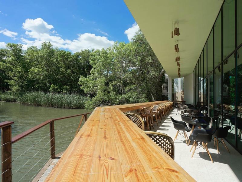 Taras z drewnianą balustradą i stolikami nad brzegiem rzeki obok nowoczesnego budynku.