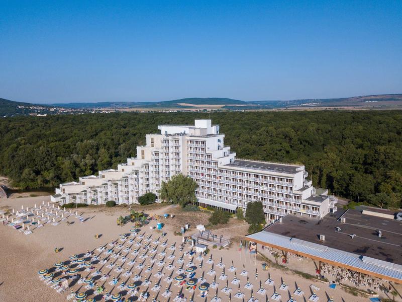 Duży hotel z tarasami, piaszczystą plażą i leżakami przed lasem pod czystym niebem.