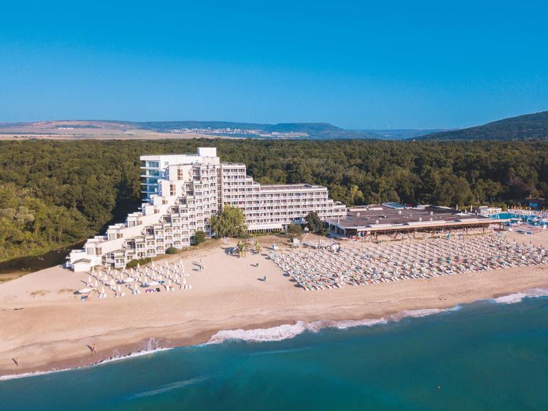 Duży hotel na piaszczystej plaży z wieloma parasolami i lasem w tle.
