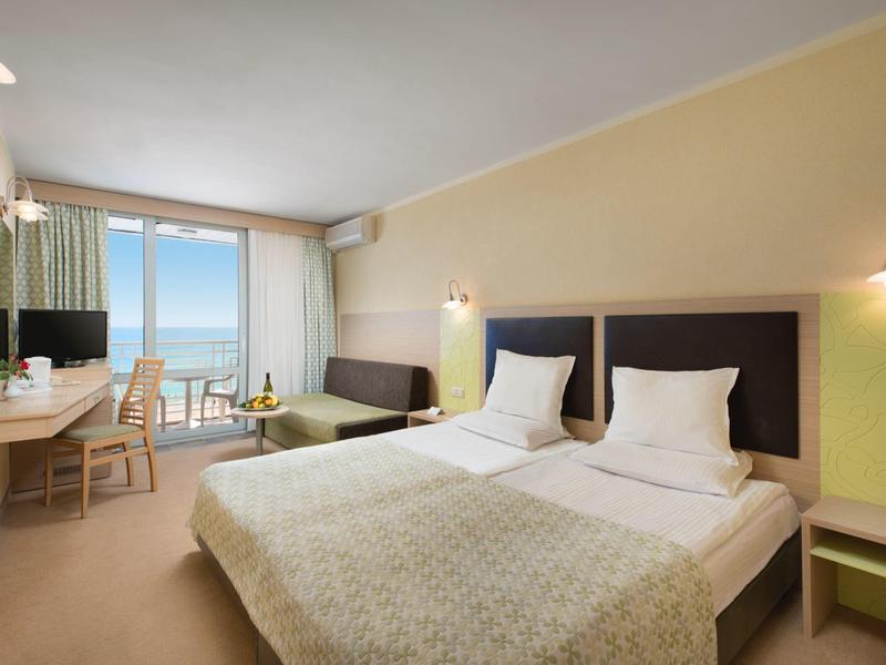 Jasny pokój hotelowy z dwoma łóżkami, biurkiem i widokiem na morze przez duże okna.