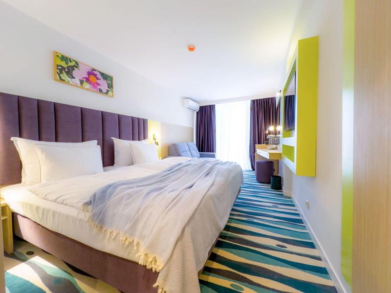 Nowoczesny pokój hotelowy z dużym łóżkiem, kolorowym dywanem i jasnym wystrojem ścian.