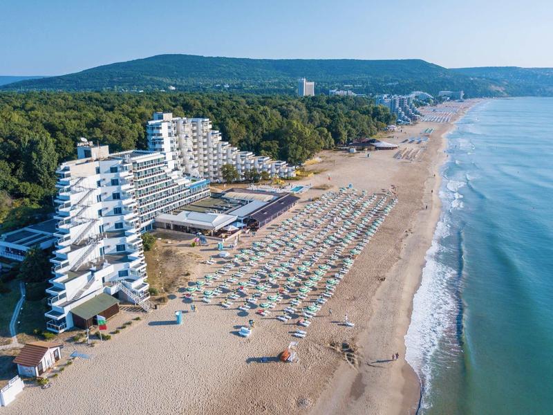 Grand hôtel sur la plage avec de nombreux parasols et vue sur les collines boisées.