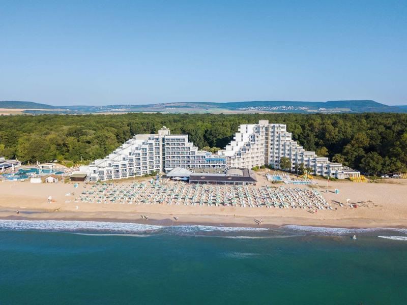 Hôtel moderne sur la plage avec parasols et forêt en arrière-plan sous un ciel clair.