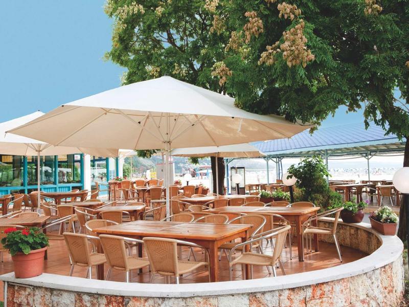 Terrazza aperta con sedie in legno, tavoli e grandi ombrelloni sotto gli alberi.