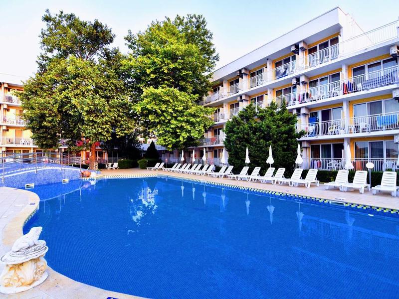 Grande piscina all'aperto con lettini accanto a un edificio dell'hotel su più piani e alberi verdi.