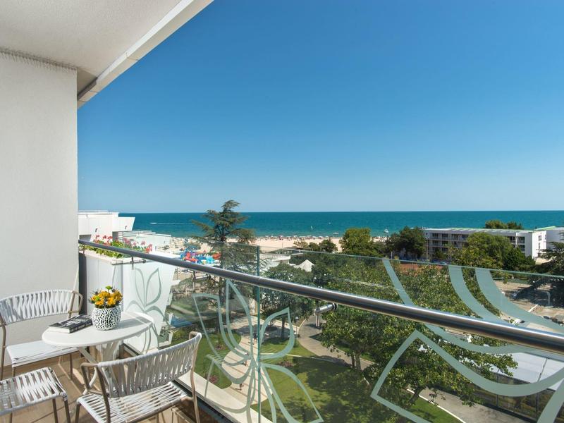 Balkon s židlemi a stolem, výhled na zahradu, pláž a moře pod jasnou oblohou.