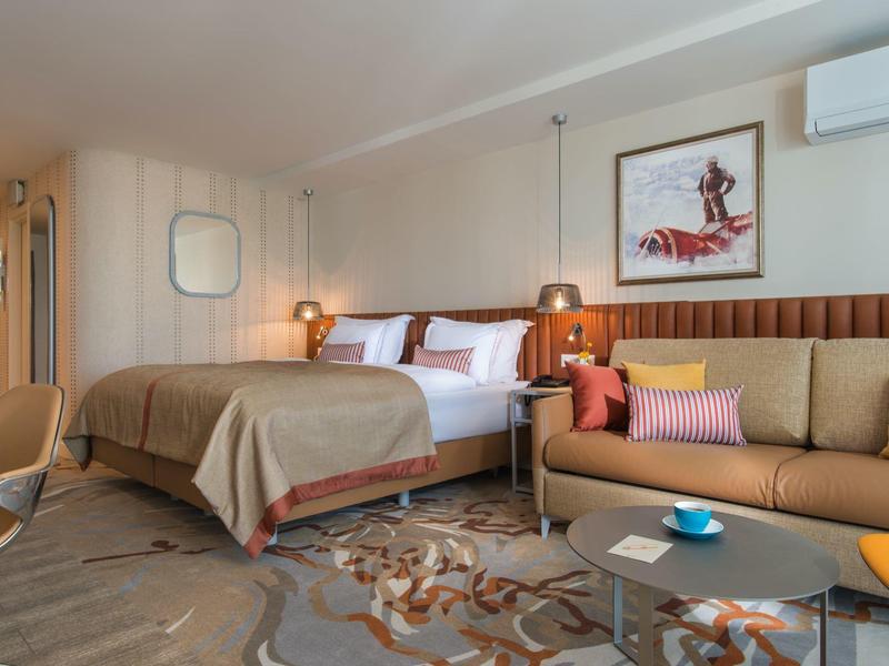 Moderní hotelový pokoj s postelí, pohovkou, kobercem a dekorativním osvětlením v teplých tónech.