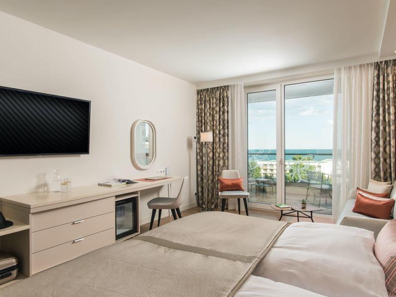 Světlý hotelový pokoj se dvěma postelemi, TV, psacím stolem a balkonem s výhledem na moře.