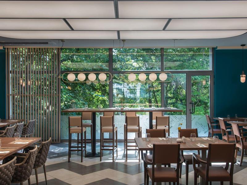 Moderní restaurace s velkými okny a výhledem na zelené stromy.