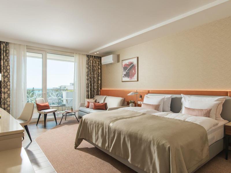 Světle zařízený hotelový pokoj s velkou postelí, křesly a výhledem na město.