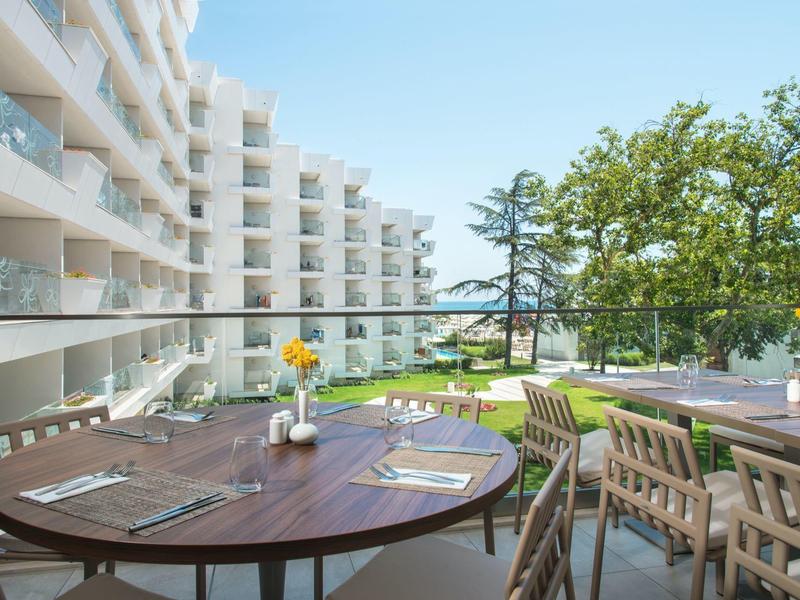 Stůl na balkoně se židlemi a výhledem na hotelovou terasu a zahradu se stromy.