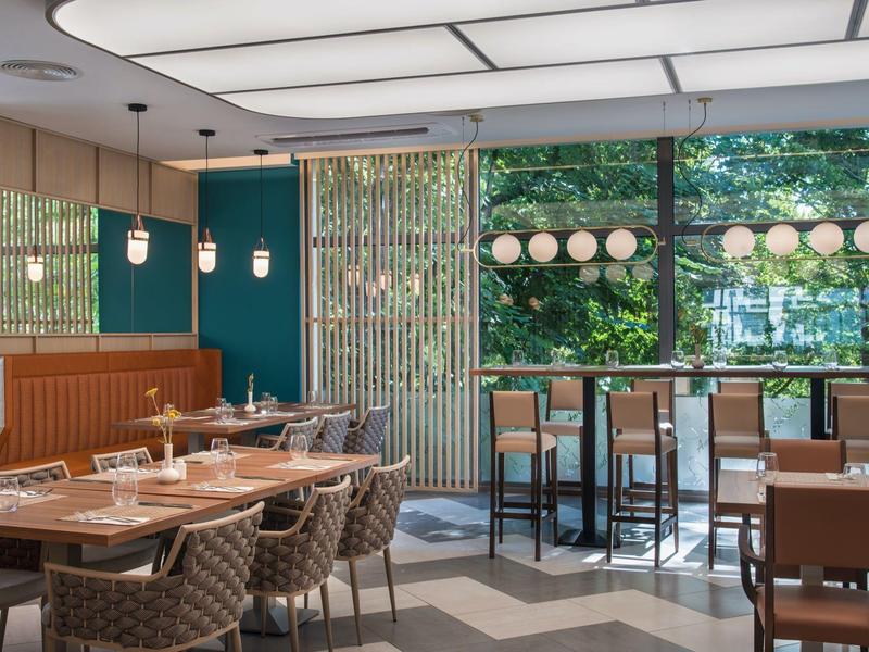 Moderní restaurace s dřevěnými stoly, čalouněnými židlemi a velkými okny s výhledem do zeleně.