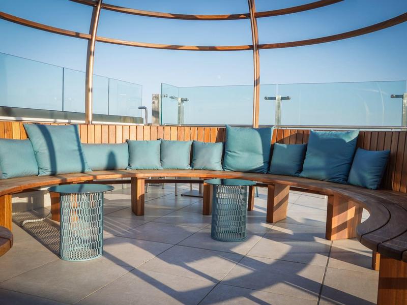 Kruhová venkovní posezení ze dřeva s modrými polštáři a skleněnými stolky na terase se skleněným zábradlím.