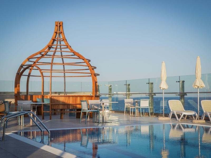 Moderní bazén na střeše hotelu s lehátky a dřevěným altánem za jasné oblohy.