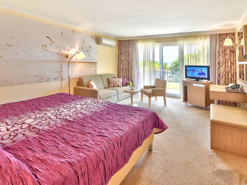 Světle zařízený hotelový pokoj s velkou postelí, posezením a balkonem s výhledem na zeleň.