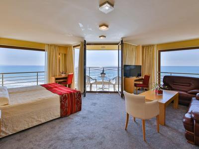 Světý hotelový pokoj s velkou postelí, posezením a výhledem na moře přes velká okna.