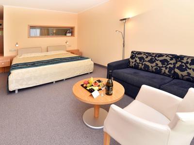 Moderní hotelový pokoj s manželskou postelí, stolem, židlemi a pohovkou v teplém osvětlení.