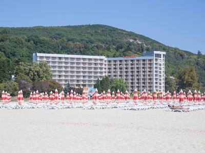 Pláž s bílými slunečníky a hotelem před zalesněným kopcem za jasné oblohy.