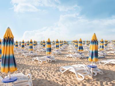 Mnoho zavřených žluto-modrých plážových slunečníků a bílých lehátek na písčité pláži pod modrou oblohou.