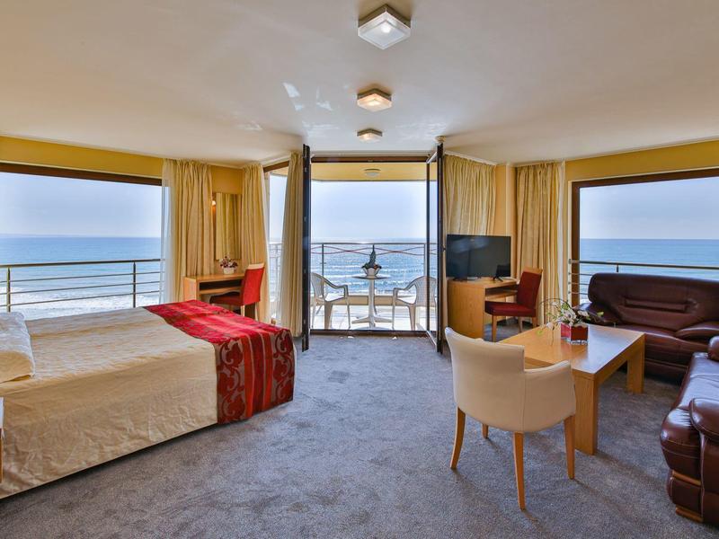 Světý hotelový pokoj s velkou postelí, posezením a výhledem na moře přes velká okna.