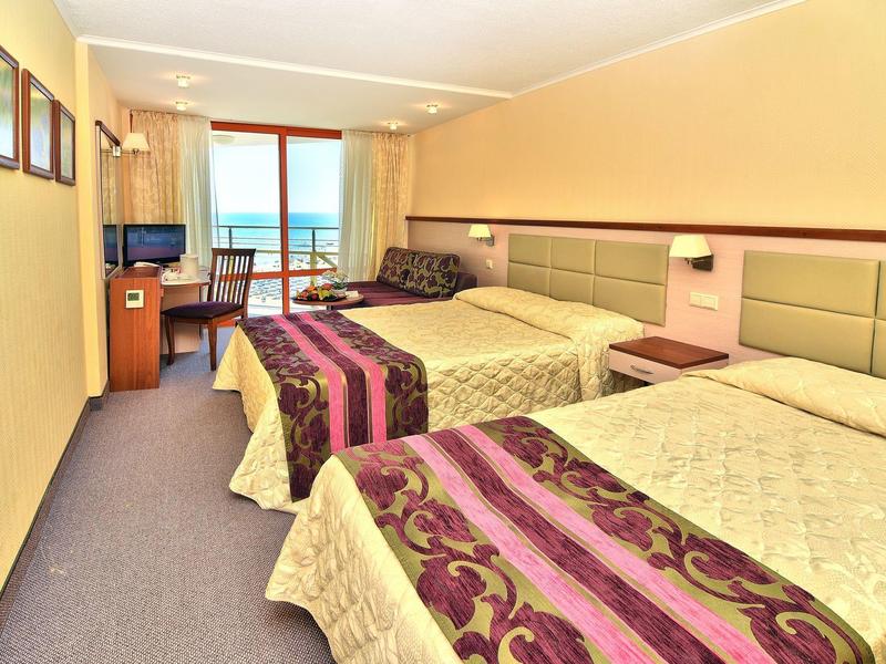 Světý hotelový pokoj se dvěma dvojlůžky, psacím stolem, TV a výhledem na moře přes balkonové dveře.