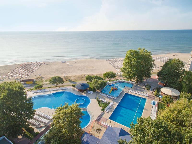 Výhled na pláž, moře, dva bazény a lehátka v hotelu obklopeném stromy.