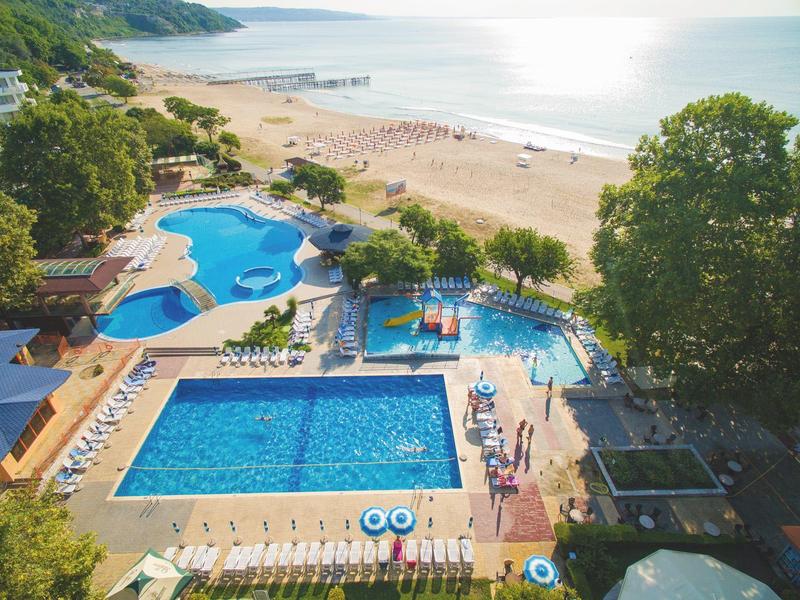 Hotelová bazénová zóna s bazény a výhledem na pláž u moře, obklopená stromy a lehátky.