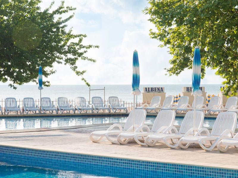 Prázdná bazénová plocha s bílými lehátky a modrými slunečníky, obklopená stromy.