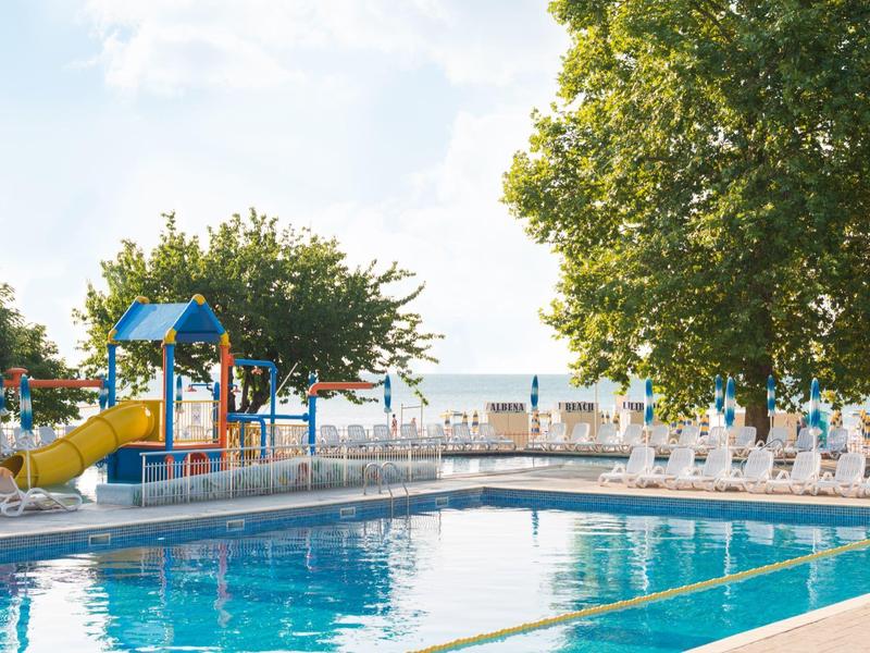 Rekreační bazén s vodní skluzavkou a lehátky pod velkými stromy za slunečného dne