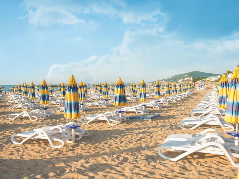 Mnoho bílých lehátek a žluto-modrých slunečníků na písečné pláži pod modrou oblohou