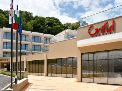 Modernes, mehrstöckiges Hotelgebäude mit großen Fenstern, drei Flaggen und grünem Wald im Hintergrund.