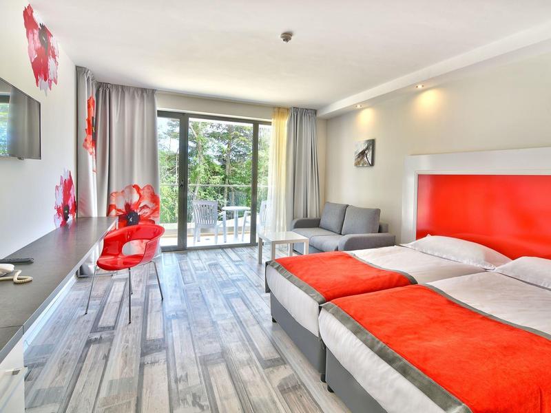 Helles Hotelzimmer mit Doppelbett, roter Einrichtung, Balkon und modernem Holzfußboden.