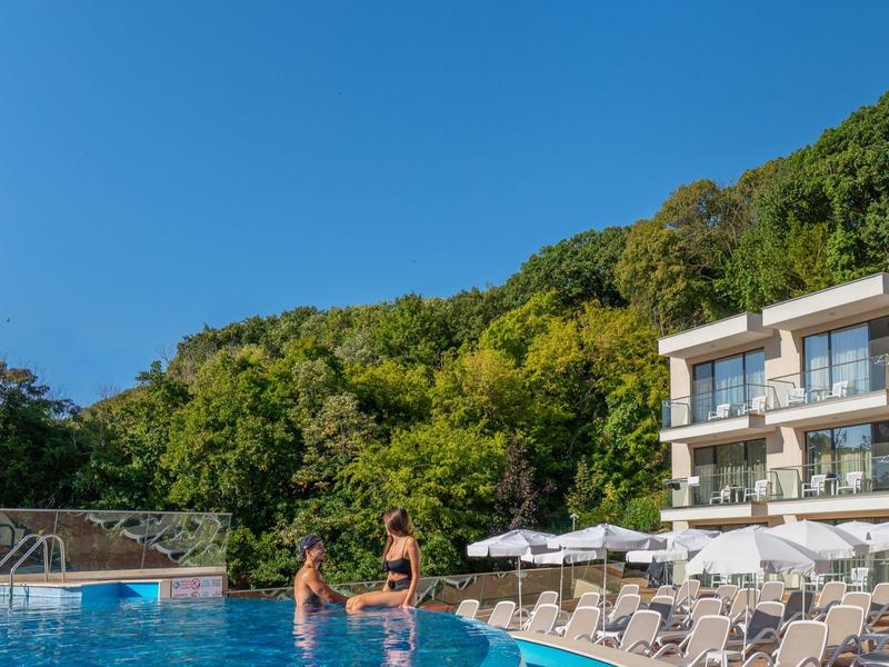 Zwei Personen entspannen am Pool eines Hotels mit Liegestühlen und grünem Wald im Hintergrund.