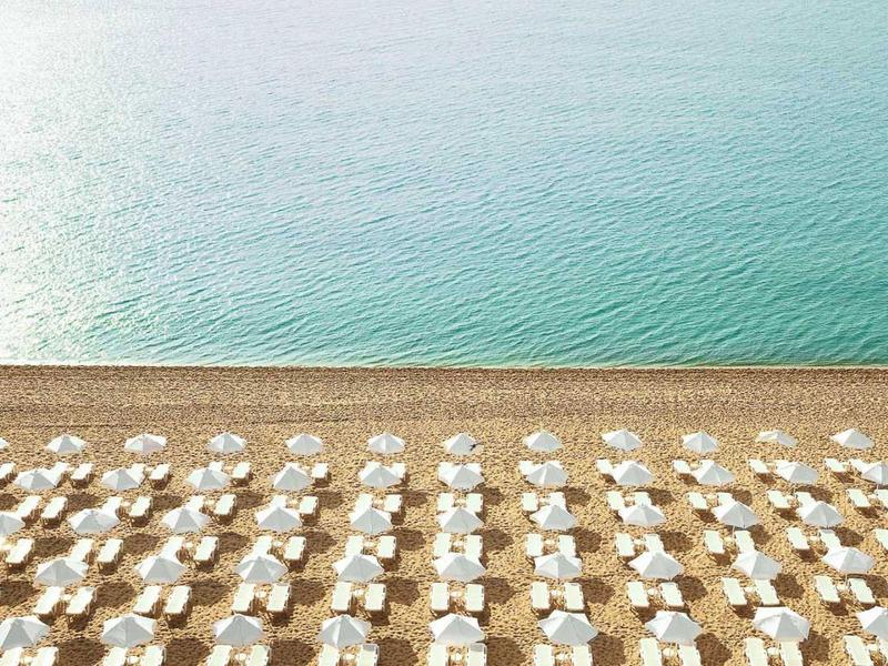Symmetrisch angeordnete Sonnenschirme und Liegestühle am Strand mit ruhigem Meer im Hintergrund.