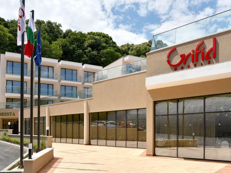 Modernes, mehrstöckiges Hotelgebäude mit großen Fenstern, drei Flaggen und grünem Wald im Hintergrund.