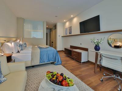 Moderní hotelový pokoj s manželskou postelí, pracovním stolem, televizí a kancelářskou židlí.