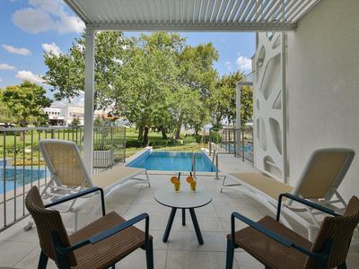 Balkon s posezením s výhledem na bazén a stromy pod modrou oblohou.