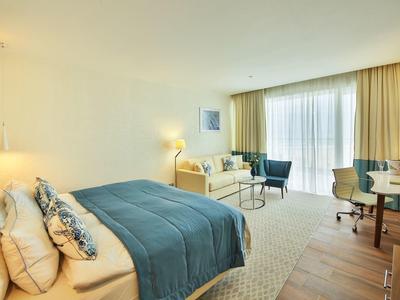 Světlý hotelový pokoj s manželskou postelí, modrým přehozem, posezením a velkým oknem se záclonami.