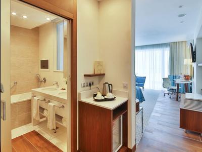 Moderní hotelová chodba s dřevěnou podlahou, pohled na koupelnu a pokoj s posezením.