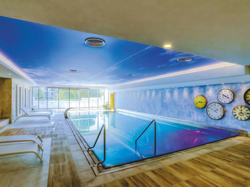 Vnitřní bazén s dřevěnou podlahou, nástěnnými hodinami a lehátky v moderním wellness.