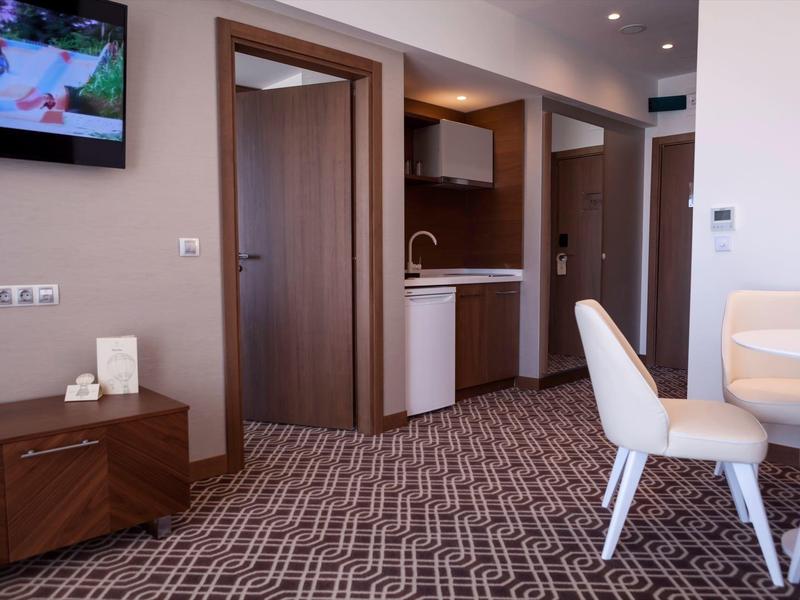 Moderní hotelový pokoj s kobercem, kuchyňským koutem, židlemi a televizí.